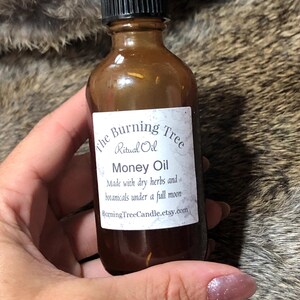 May include: A brown glass bottle with a black lid, labeled "The Burning Tree Ritual Oil Money Oil". The label also states "Made with dry herbs and botanicals under a full moon" and "BurningTreeCandle.etsy.com".