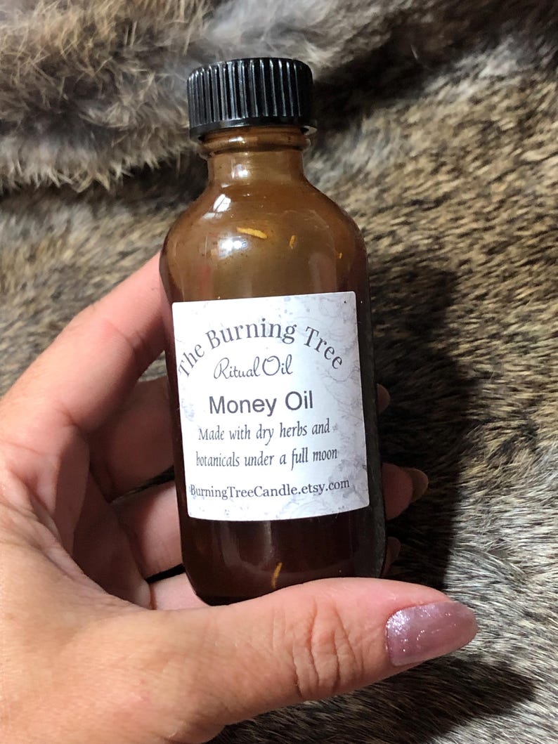 May include: A brown glass bottle with a black lid, labeled "The Burning Tree Ritual Oil Money Oil". The label also states "Made with dry herbs and botanicals under a full moon" and "BurningTreeCandle.etsy.com".