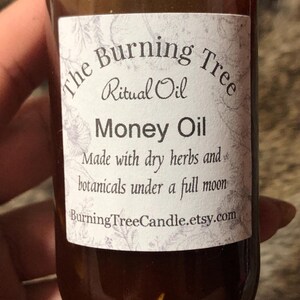 May include: A brown glass bottle with a white label that reads "The Burning Tree Ritual Oil Money Oil Made with dry herbs and botanicals under a full moon BurningTreeCandle.etsy.com"