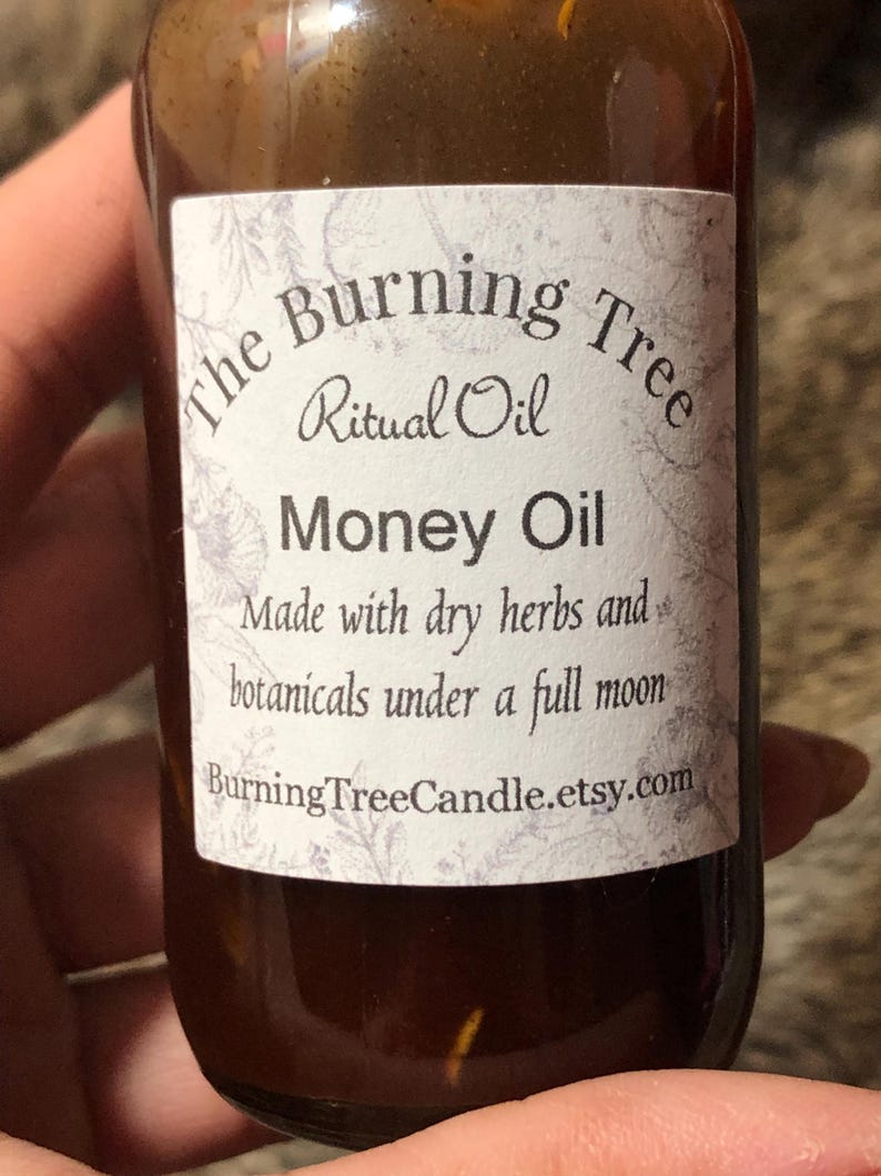 May include: A brown glass bottle with a white label that reads "The Burning Tree Ritual Oil Money Oil Made with dry herbs and botanicals under a full moon BurningTreeCandle.etsy.com"