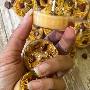 Puede incluir: Primer plano de una mano sosteniendo una vela peque&ntilde;a, redonda, de color naranja con flores secas y piedras marrones incrustadas en la cera. La vela est&aacute; rodeada de otras velas similares.