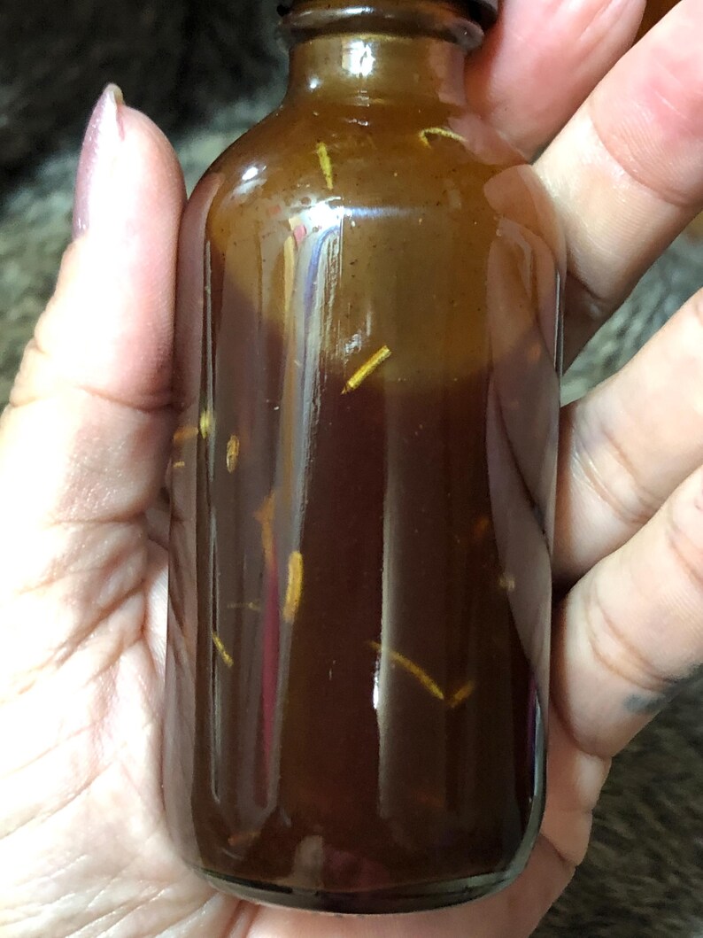 May include: A small, amber glass bottle filled with a dark brown liquid and containing small, light brown pieces. The bottle has a screw-on lid.