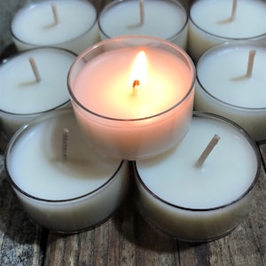 May include: A group of eight white tea light candles, one of which is lit, arranged in a circle on a wooden surface.