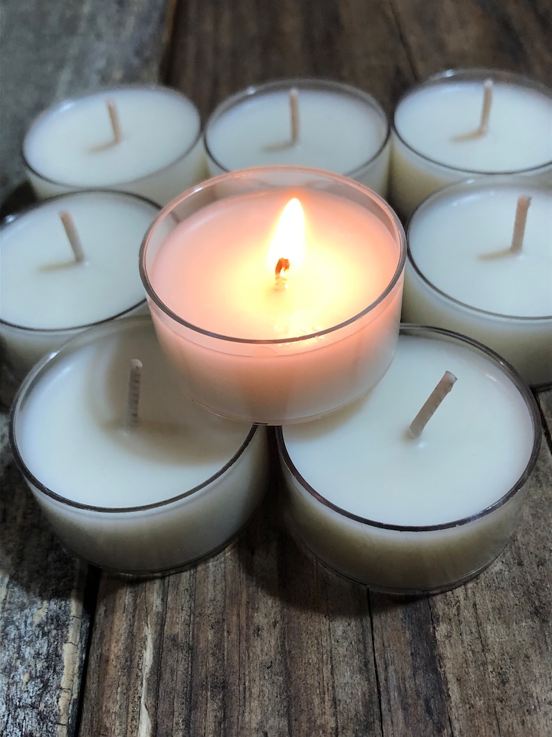 May include: A group of eight white tea light candles, one of which is lit, arranged in a circle on a wooden surface.