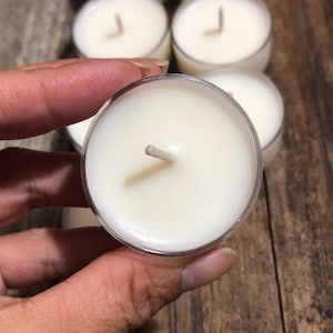 May include: A close-up of a white tea light candle with a wick in a clear plastic container. The candle is being held by a hand.