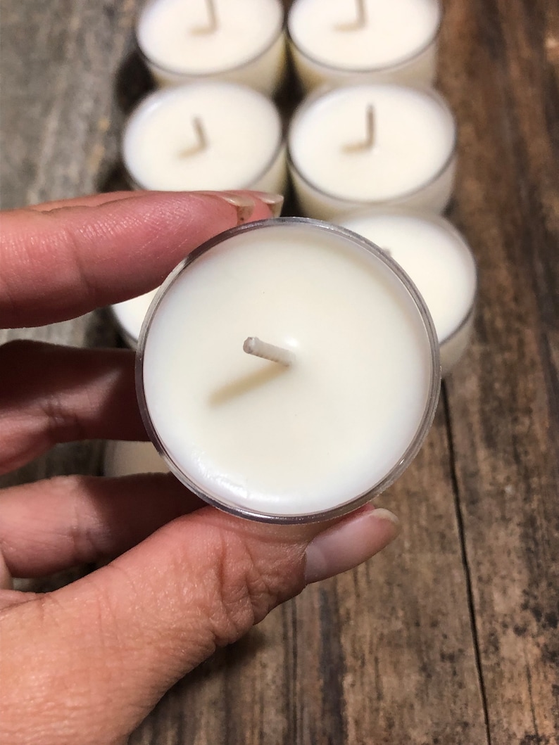 May include: A close-up of a white tea light candle with a wick in a clear plastic container. The candle is being held by a hand.