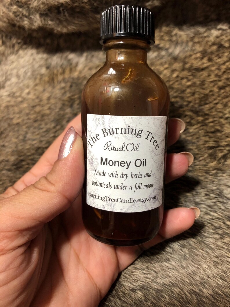 May include: A small brown glass bottle with a black lid, labeled "The Burning Tree Ritual Oil Money Oil" and "Made with dry herbs and botanicals under a full moon" with the website "BurningTreeCandle.etsy.com".