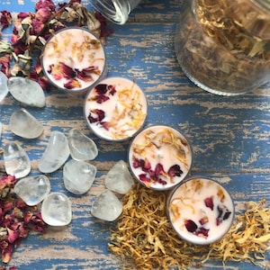 May include: Four pink tea light candles with dried flower petals and a scattering of dried flowers on a blue wooden surface. There are also clear quartz crystals and a jar of dried flowers.