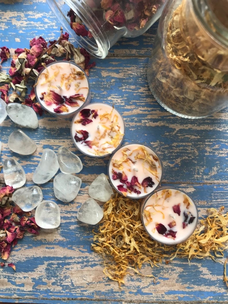 May include: Four pink tea light candles with dried flower petals and a scattering of dried flowers on a blue wooden surface. There are also clear quartz crystals and a jar of dried flowers.