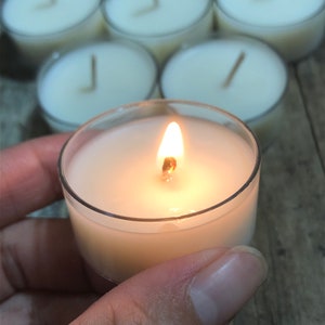 May include: A single white tea light candle burning in a clear glass holder. The candle is surrounded by other unlit tea lights.