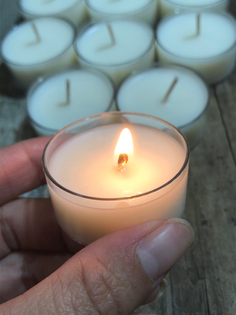 May include: A single white tea light candle burning in a clear glass holder. The candle is surrounded by other unlit tea lights.