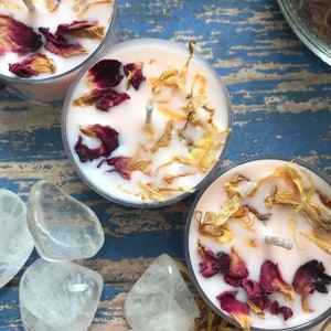 May include: Three pink tea light candles with dried flower petals and a white wick. The candles are on a blue and white wooden surface with clear quartz stones.