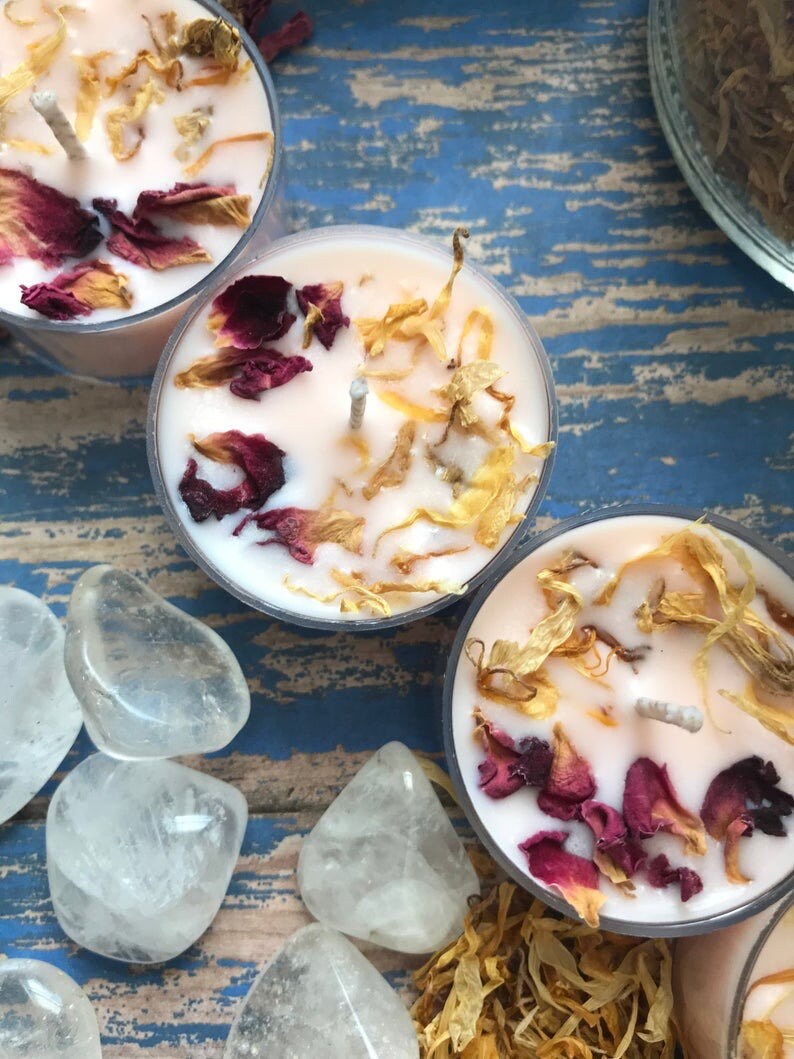 May include: Three pink tea light candles with dried flower petals and a white wick. The candles are on a blue and white wooden surface with clear quartz stones.