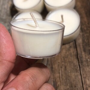 May include: A close-up of a white tea light candle in a clear plastic container. The candle has a single wick and is surrounded by other tea light candles.