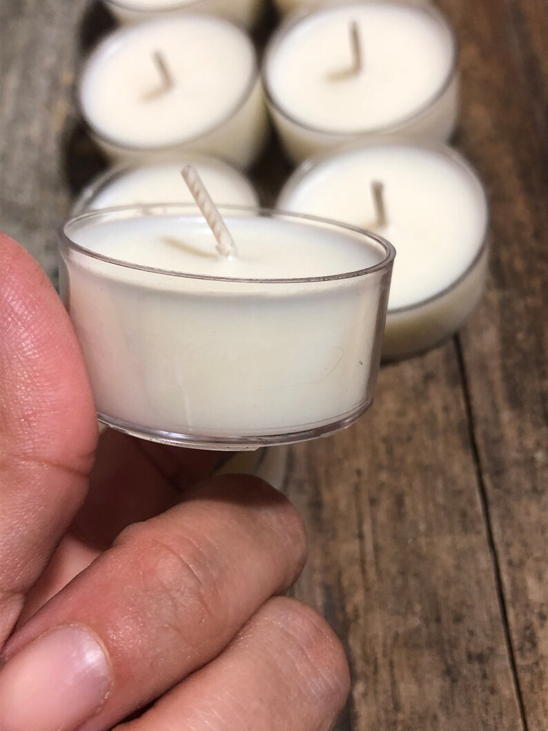 May include: A close-up of a white tea light candle in a clear plastic container. The candle has a single wick and is surrounded by other tea light candles.