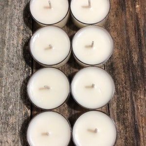 May include: Eight white unscented tea light candles in clear plastic containers arranged in two rows of four on a wooden surface.