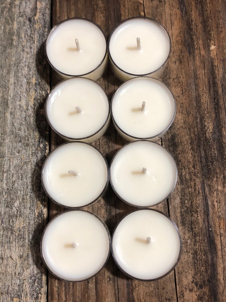 May include: Eight white unscented tea light candles in clear plastic containers arranged in two rows of four on a wooden surface.