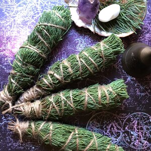 May include: Four bundles of dried cedar sage smudge sticks tied with twine. The bundles are arranged on a dark purple background with a map pattern.