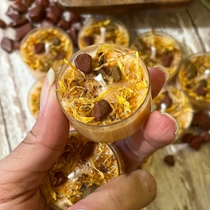 Puede incluir: Primer plano de una mano sosteniendo una vela peque&ntilde;a, redonda, marr&oacute;n y amarilla con p&eacute;talos de flores secas y peque&ntilde;as piedras marrones incrustadas en la cera. La vela est&aacute; rodeada de otras velas similares.