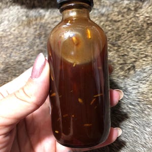 May include: A small amber glass bottle with a black screw-on lid filled with a dark brown liquid and small, light brown particles.