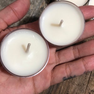 May include: Two white tea light candles with wicks in clear plastic containers. The candles are sitting in a hand.