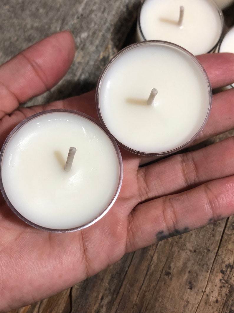 May include: Two white tea light candles with wicks in clear plastic containers. The candles are sitting in a hand.