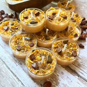 Puede incluir: Una colecci&oacute;n de peque&ntilde;as velas de t&eacute; redondas de color naranja con p&eacute;talos de flores secas y peque&ntilde;as piedras incrustadas en la cera. Las velas est&aacute;n dispuestas en c&iacute;rculo sobre una superficie de madera blanca.