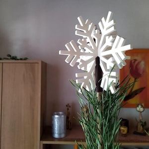 Könnte beinhalten: Ein weißer Weihnachtsbaumschmuck in Form einer Holz-Schneeflocke. Die Schneeflocke hat ein detailliertes, mehrschichtiges Design. Der Aufsatz ist auf einem braunen Pfahl montiert, umgeben von grünem Gras.
