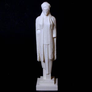 Könnte beinhalten: Eine cremefarbene Statue einer bekleideten Figur, die auf einem rechteckigen Sockel steht. Die Figur hat ein drapiertes Gewand und einen ruhigen Ausdruck. Die Statue steht vor einem schwarzen Hintergrund.