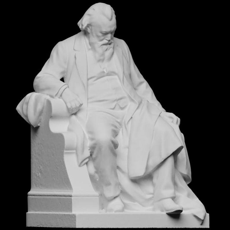 Op de afbeelding: Een witte sculptuur van een zittende man met een lange baard, gekleed in een pak en jas. De figuur zit op een stoel met een hoge rugleuning en kijkt naar beneden. De sculptuur staat tegen een zwarte achtergrond.