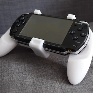 Może przedstawiać: Biały plastikowy uchwyt do gier ręcznych dla czarnej konsoli Sony PlayStation Portable (PSP). Kontroler ma dwa białe uchwyty po bokach i biały zaczep, który utrzymuje PSP w miejscu.
