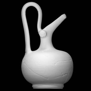 Può includere: Una brocca in ceramica bianca con un manico curvo e un beccuccio. La brocca ha un corpo bulboso con crepe visibili e un elemento circolare decorativo dove il manico si collega al corpo. Lo sfondo è nero.