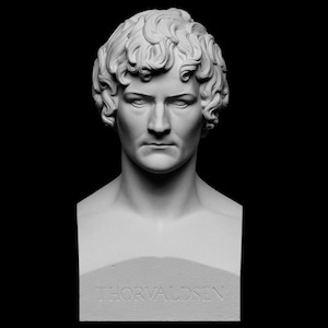 Op de afbeelding: Een witte buste van een man met krullend haar en een serieuze uitdrukking. De buste staat op een sokkel met de naam "THORVALDSEN" erin gegraveerd. De achtergrond is zwart.