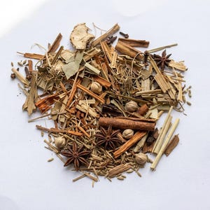 Op de afbeelding: Aromatische mix van gedroogde kruiden en specerijen, waaronder steranijs, kaneelstokjes, kardemompeulen en gember. De mix heeft verschillende tinten bruin, beige en groen. De ingrediënten zijn verspreid op een witte ondergrond.