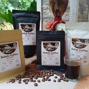 Puede incluir: Una variedad de productos de café, incluyendo bolsas de café Suluk, granos enteros y un vaso de café. Un molinillo de café rojo es visible en el fondo. Las bolsas son negras, blancas y beige.