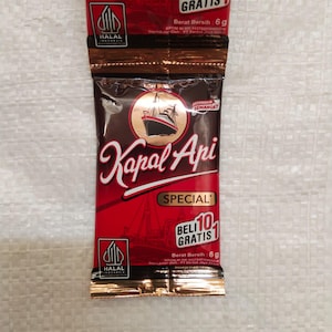 Könnte beinhalten: Zwei rot-braune Kaffeepäckchen mit dem Text "Kapal Api" in weißer Schrift. Die Päckchen haben ein Schiffslogo und die Worte "Special" und "Beli 10 Gratis 1". Jedes Päckchen enthält 6g Kaffee und hat ein Halal-Logo.