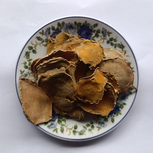 May include: A white ceramic plate with a blue floral border holds a pile of dried, sliced turmeric. The turmeric slices are various shades of brown and orange. The plate is on a white surface.