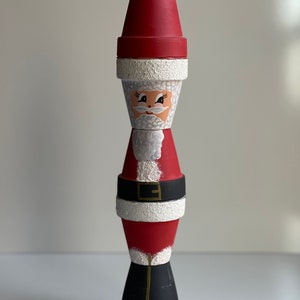 May include: A hand-painted Santa Claus figurine made from stacked terracotta pots. The figurine is decorated with red, white, and black paint, and features a white cotton ball for a hat.