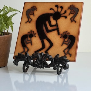 May include: A brown ceramic tile with a black silhouette of a Native American flute player and dancers. The tile is on a black metal stand.