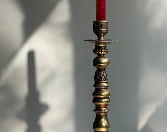 May include: A gold metal candle holder with ornate details and a red candle. The candle is lit and the holder is on a white surface.