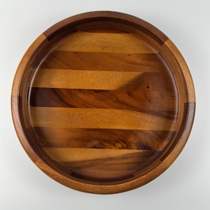 May include: A round wooden serving bowl with a dark brown rim and a lighter brown interior. The bowl is made of several wooden planks arranged in a circular pattern. The bowl is empty and sits on a white surface.
