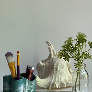 May include: A green and white ceramic makeup brush holder with three compartments. Two brushes and a lipstick are in the holder. A white seashell and a glass vase with white stones and a green plant are in the background.