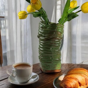May include: A green ceramic vase with a textured surface holds a bouquet of yellow tulips. A white coffee cup with a brown liquid sits on a wooden table next to a croissant on a gray plate.