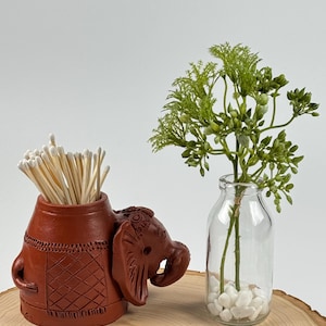 Peut inclure: Un porte-allumettes en forme d'éléphant en terre cuite avec un motif décoratif. Le porte-allumettes est rempli d'allumettes en bois et est posé sur une surface en bois. Un petit vase en verre avec des pierres blanches et une plante verte se trouve à côté du porte-allumettes.