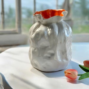 May include: White ceramic vase with an orange rim and a textured, organic shape. The vase is sitting on a white table with a pink tulip.