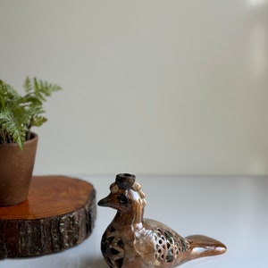 May include: A brown ceramic bird figurine with a detailed design and a small hole on top. The bird has a brown and green glaze and a textured surface.