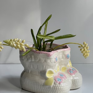 May include: White ceramic planter shaped like a baby's shoe with pink and yellow accents. The planter has a green plant with white flowers inside.