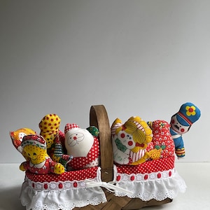 May include: A wicker basket with a white lace trim filled with colorful fabric dolls. The dolls include a Santa Claus, a girl with blonde hair, a boy in a blue uniform, and two other dolls.