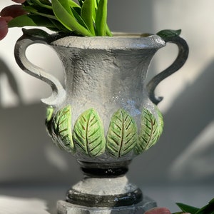 May include: A gray ceramic vase with green leaf details and two handles. The vase is filled with pink tulips.