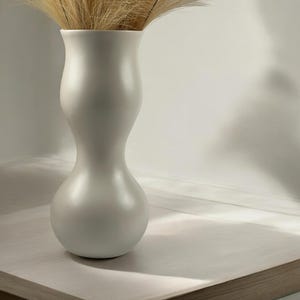 May include: A white ceramic vase with a bulbous shape holds a bouquet of dried beige pampas grass. The vase sits on a light wood surface with a darker wood border.